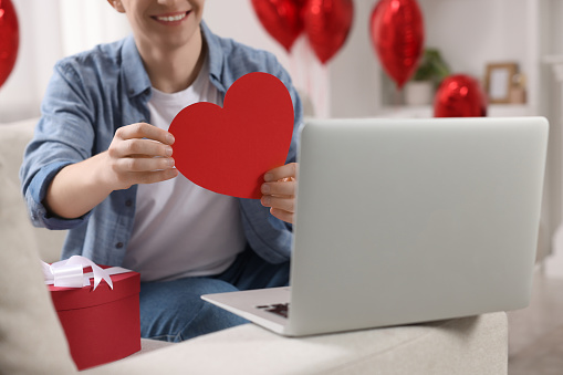 Valentine's day celebration in long distance relationship. Man holding red paper heart while having video chat with his girlfriend via laptop, closeup