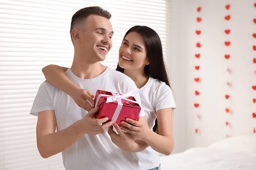 Girlfriend presenting gift to her boyfriend indoors, space for text. Valentine`s day celebration