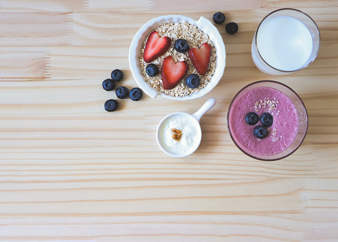 Top view or flat lay of  breakfast with oat or granola in white bowl, fresh blueberries, strawberries, a  glass of milk, smoothie blueberries  and yogurt on wooden table. Healthy breakfast concept.