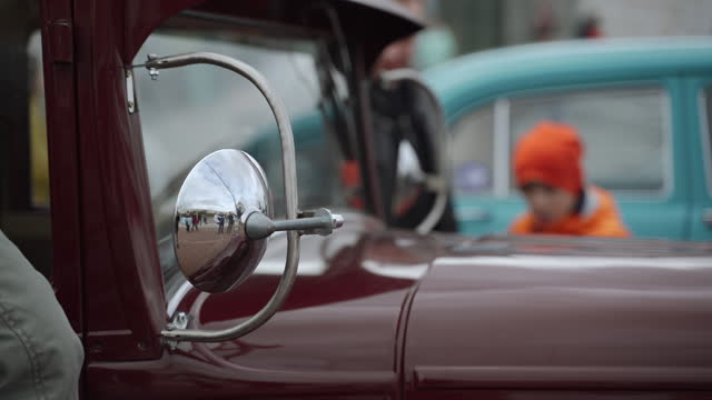 Windshield with chrome mirrors of ancient vintage car on display with people.