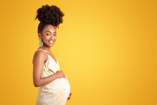 Happy young african american pregnant woman in dress touching big belly with hand, isolated on yellow studio background. Expectation child, health care, motherhood, ad and offer