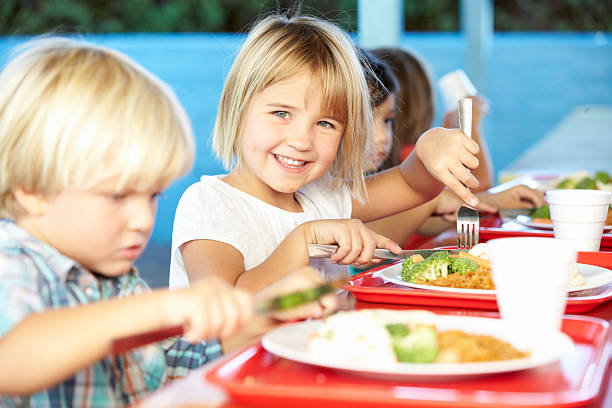 Elementary Pupils Enjoying Healthy Lunch In Cafeteria Elementary Pupils Enjoying Healthy Lunch In Cafeteria Sitting Down Smiling At Camera school lunch child food lunch stock pictures, royalty-free photos & images