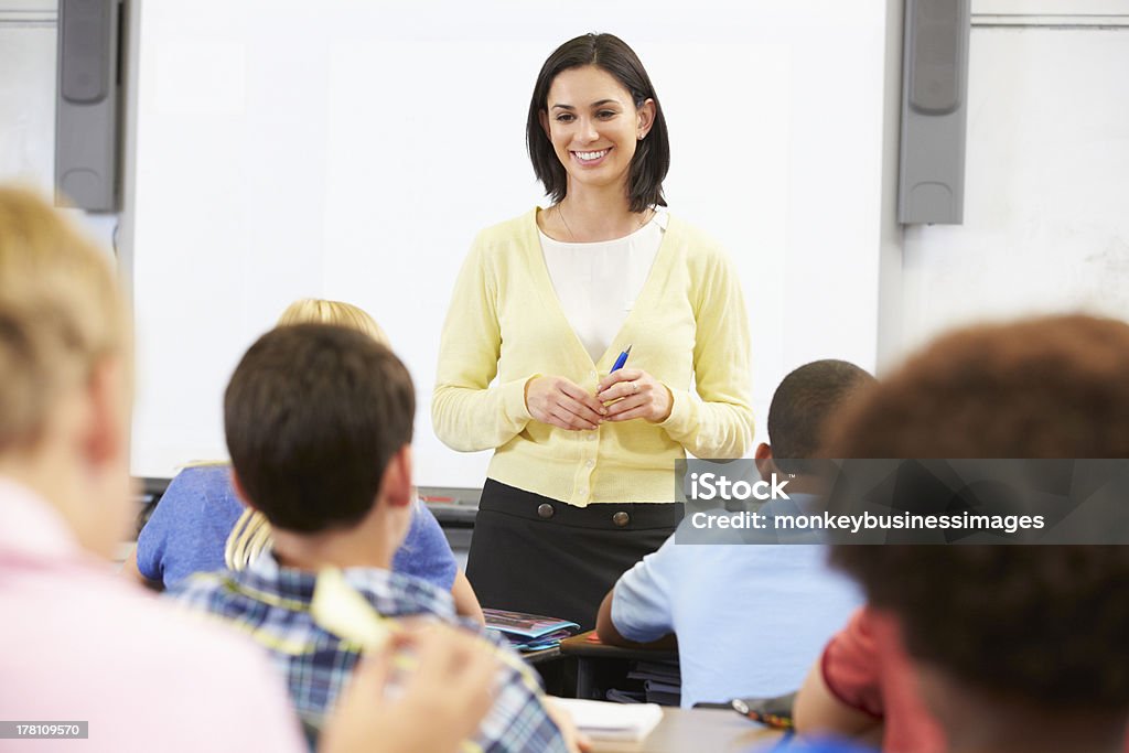 Female teacher holding pen while standing in front of class Teacher Standing In Front Of Class Of Pupils Looking At Them Smiling Teacher Stock Photo
