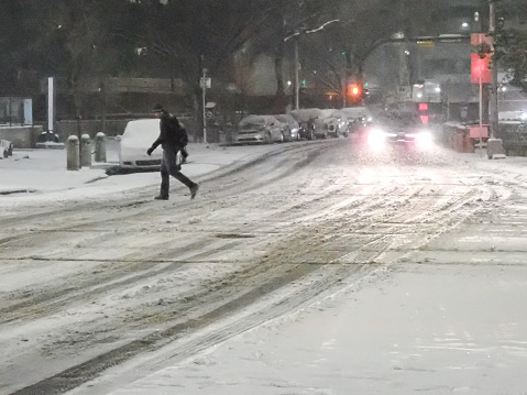 A pedestrian crossing a snow-covered street at night with the headlights of a car illuminating the snowfall.  Calgary, Alberta, Canada.
