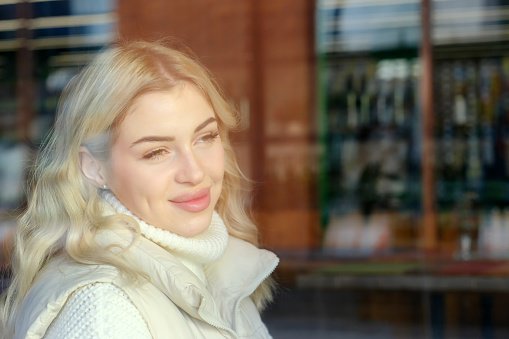 Portrait of a young beautiful blonde with long hair in a white sweater and jacket resting in a cafe, view from the street through the window