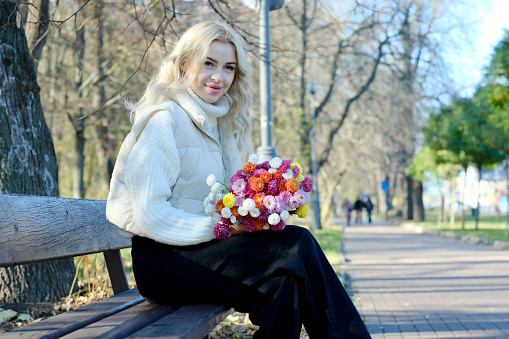 Portrait of a young beautiful blonde with long hair in a white sweater and jacket in sunny autumn in a city park with a bouquet of flowers