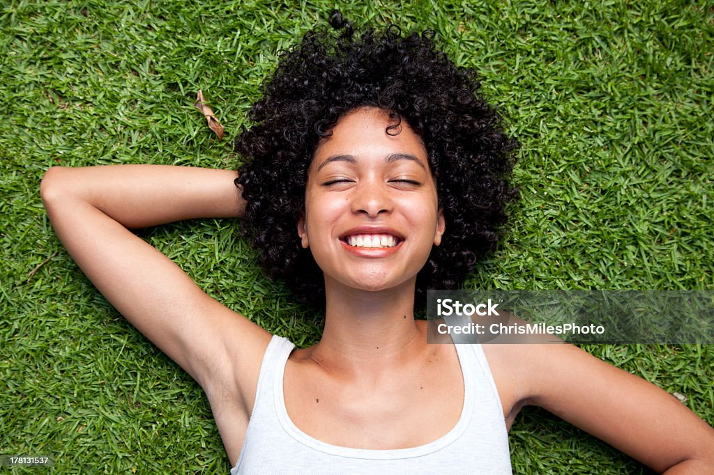Beautiful Woman Lying Down on Grass Happy mixed race woman lying down on green grass with hand behind her head. Women Stock Photo