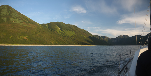 Approaching wild uninhabited coast with mountains on sail yacht, ship side visible, panoramic view, entering Morzhovaya bay, famous Kamchatka travel destination