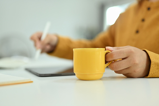 Cropped shot of woman hand holding cup of coffee and writing notes on digital tablet.