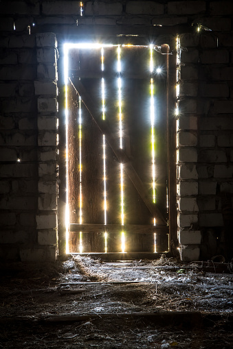 Rays of light shine through the plank door of an old barn. An old door in a brick doorway.