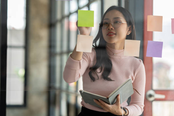 istock Confident businesswoman in eyeglasses using a notebook to jot down ideas Idea goes on post-it note on glass wall for strategic business plan, develop organization to success. 1781432849