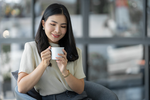Attractive young Asian woman or businesswoman sitting drinking and holding cup of coffee relaxing, taking break while sitting cross legged on sofa chair at office.