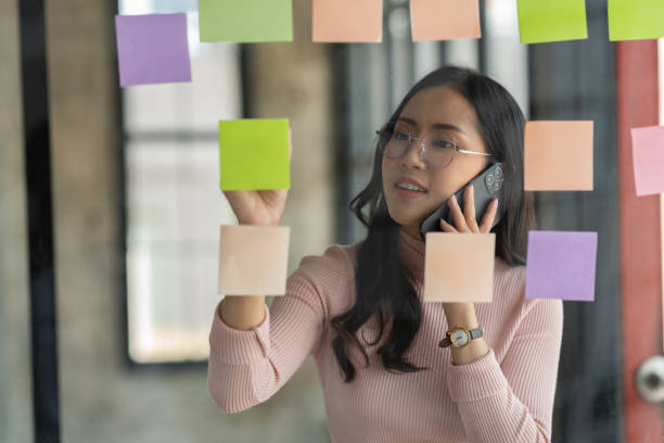 Confident businesswoman in eyeglasses using mobile phone to communicate and discuss ideas on post-it notes on glass wall for strategic business planning, organizational development to success. mujer de negocios segura de sí misma en anteojos que usa el teléfono móvil para comunicarse y discutir ideas en notas adhesivas en la pared de vidrio para la planificación estratégica del negocio, el desarrollo organizacional para el éxito.
