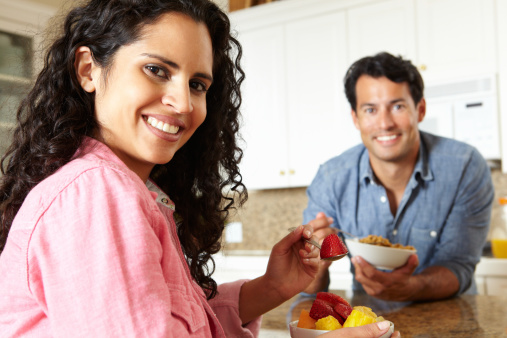 Hispanic couple eating cereal and fruit sitting at kitchen table smiling at camera