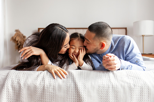 happy young asian family with little daughter lie on bed at home and smile, korean girl together with parents rest at home on bed, mom and dad kiss and love daughter