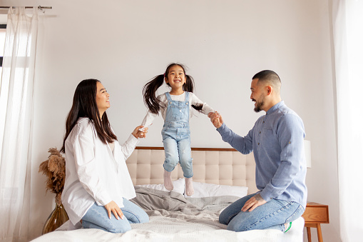 happy asian family with daughter play and rejoice at home on the bed, little korean girl jumping on the bed and holding hands mom and dad
