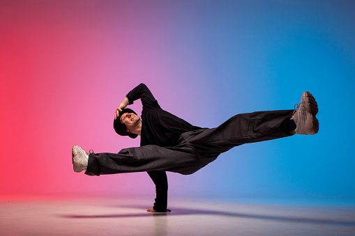 young guy dancer is dancing break in neon lighting, male acrobat is doing trick and dance exercise on red blue background, hip hop performer is moving and standing on his hands