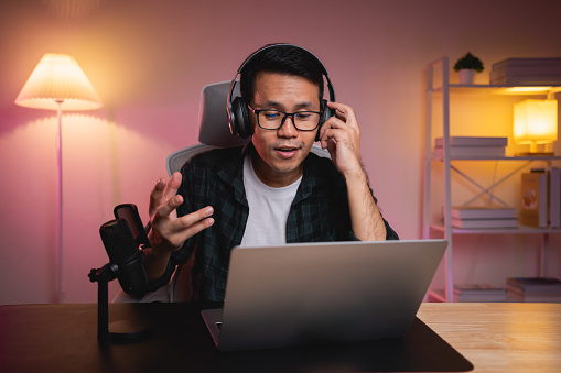 Asian influencer man wearing glasses smile youtuber using laptop live streaming greeting video conference with laptop and wearing headphone. Asian man teaching online concept.