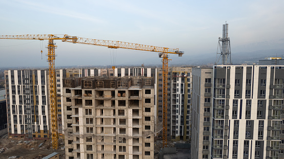 Construction of tall houses near the road. The city of Almaty. New multi-storey buildings are being built. Builders, crane, machines are working. Aerial view of houses and roads from the drone.