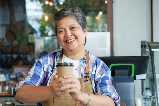 Retired Asian business woman with gray hair Standing in front holding disposable brown coffee cup bright smile. Standing front cash register counter take promotional photos for small family cafe.