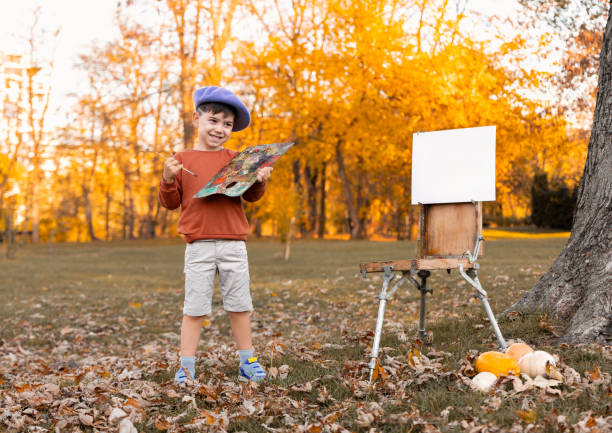 boy kid is painting in park on old easel using oil colors. child with beret and brush against autumn golden landscape.pumpkins as decoration.