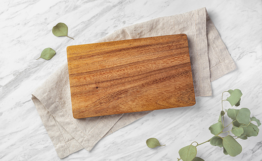Wood board on linen napkin on marble kitchen table wtih green leaves, top view