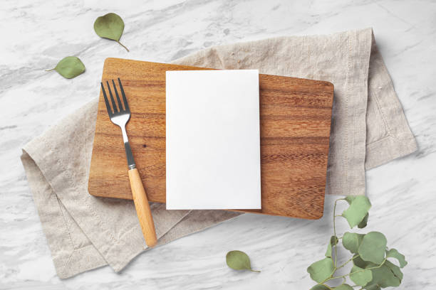 Wooden board and menu card mockup with fork and linen napkin on marble kitchen table wtih green leaves Wooden board and menu card mockup with fork and linen napkin on marble kitchen table wtih green leaves, top view restaurant table card stock pictures, royalty-free photos & images
