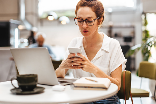 Focused female business professional checks her mobile phone for messages as she works remotely from a cozy coffee shop, managing her work tasks and communications with ease.