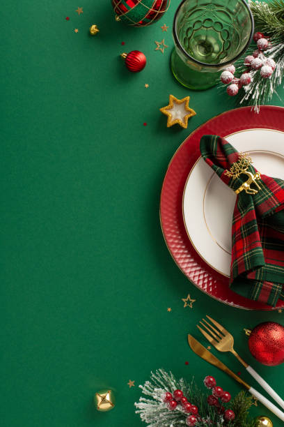 istock Compose a festive dining table layout. Top view vertical picture of plates, napkin ring, wineglass, ornaments, confetti, frosty fir branches, mistletoe, green background with space for text or ad 1781640976