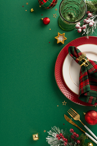 Compose a festive dining table layout. Top view vertical picture of plates, napkin ring, wineglass, ornaments, confetti, frosty fir branches, mistletoe, green background with space for text or ad