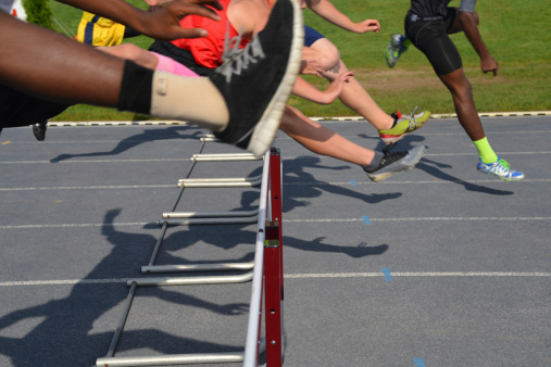 Athletes jumping over hurdles, in midair, with shadows.