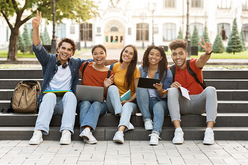 College Years Concept. Group Of Happy Multiethnic Students Posing Outdoors Together, Cheerful Young Men And Women Preparing For Exams Outside, Laughing And Showing Peace Gesture At Camera