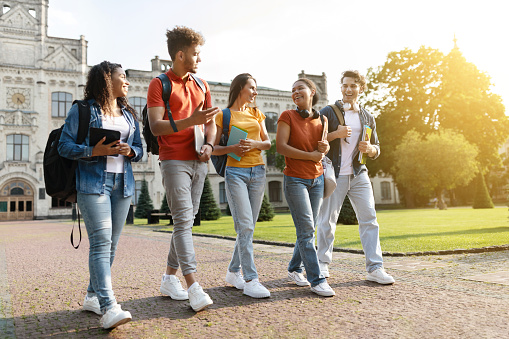 Five multiethnic students chatting and laughing while walking together at university campus, group of happy young people with books and backpacks going to classes or having fun after lessons