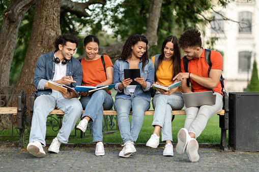 Group of multiethnic students study together outdoors in university campus, happy young college friends sitting on bench outside, preparing for exams, using digital tablet, laptop and workbooks