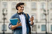 Portrait of cheerful young student guy with smartphone and workbooks posing outdoors
