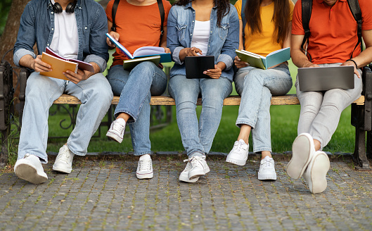 Learning Concept. Group Of Students Preparing For Exam Outdoors, Young College Friends Studying With Diverse Gadgets And Workbooks While Sitting On Bench In Campus Park, Cropped Image