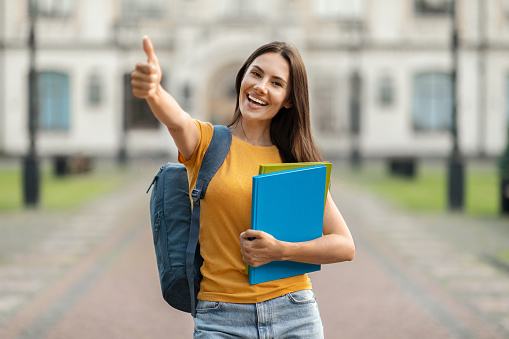 Portrait Of Beautiful Female Student Showing Thumb Up While Standing Outdoors Near University Building, Smiling Young Woman Holding Workbooks, Recommending Educational Programs, Copy Space