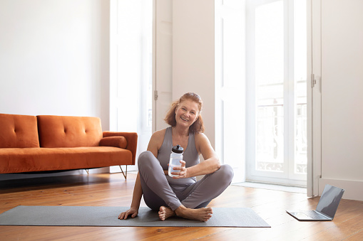 Smiling Sporty Senior Woman Relaxing After Fitness Training With Laptop At Home, Happy Older Female Sitting On Yoga Mat And Holding Water Bottle, Resting After Domestic Workout, Copy Space