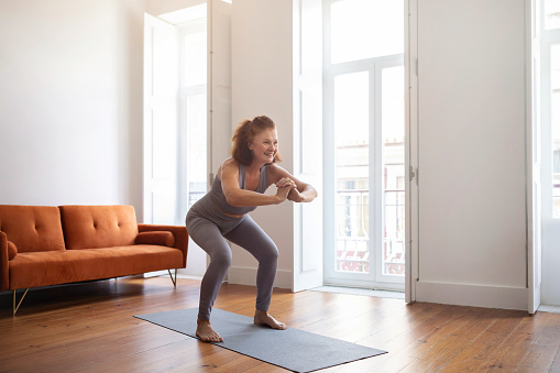 Home Workout. Smiling Senior Woman Doing Squats While Training In Living Room, Sporty Elderly Lady Exercising On Fitness Mat, Older Female Making Weight Loss Exercises, Enjoying Active Lifestyle