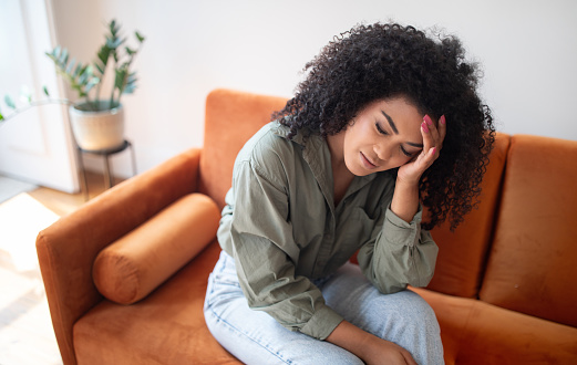 Upset black young lady suffers from headache sits on sofa touches her forehead at home. Stressed millennial woman with eyes closed having migraine health problem, feeling unwell