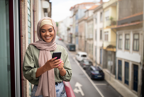 Muslim Woman In Hijab Texting On Smartphone Standing At Balcony Of Her Apartment. Happy Young Lady Using Mobile Application On New Modern Phone And Chatting In Social Media, Empty Space