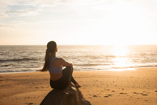 Happy calm millennial caucasian woman sit on rock, enjoy vacation, rest and peace at sea beach, sun flare, back. Morning workout, health care, sunrise and lifestyle at summer