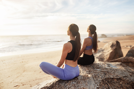 Calm glad young caucasian women enjoy morning workout, breathing exercises, meditation, practice yoga in lotus position on sea beach. Sports outdoor, peace, lifestyle and body care