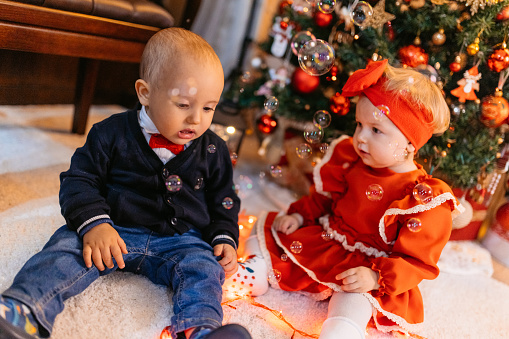 Baby boy and a baby girl sitting on the floor in front of the Christmas tree at home.