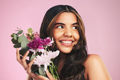 Woman, flowers and bouquet in studio for skincare, cosmetics and natural aesthetic on pink background. Face, happy model and thinking of eco beauty with floral plants, sustainability and dermatology