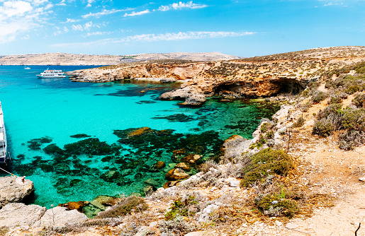 A serene view of the crystal-clear turquoise waters of the Blue lagoon of Comino island (Mediterranean Sea) in Malta. The water is surrounded by rocky cliffs, and a white boat can be seen in the distance. The sky is blue, and the vegetation is dry and sparse.