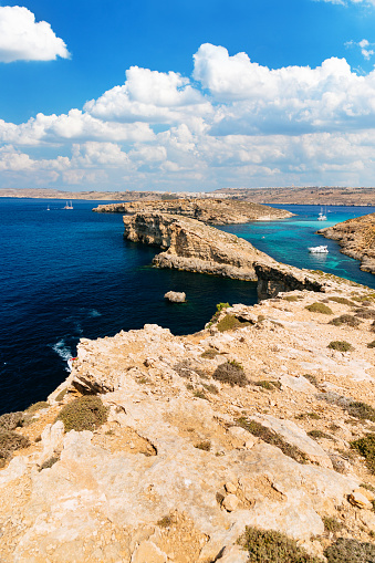 A serene view of the crystal-clear turquoise waters of the Blue lagoon of Comino island (Mediterranean Sea) in Malta. The water is surrounded by rocky cliffs, and a white boat can be seen in the distance. The sky is blue with fluffy clouds, and the vegetation is dry and sparse.