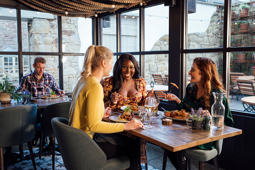 A medium wide angle view of a group of three women enjoying a catch up and dinner together on a cold winters day in a warm coastal pub in Seahouses in the North East of England. They are enjoying some pub classics and tucking in to bangers and mash and fish and chips.