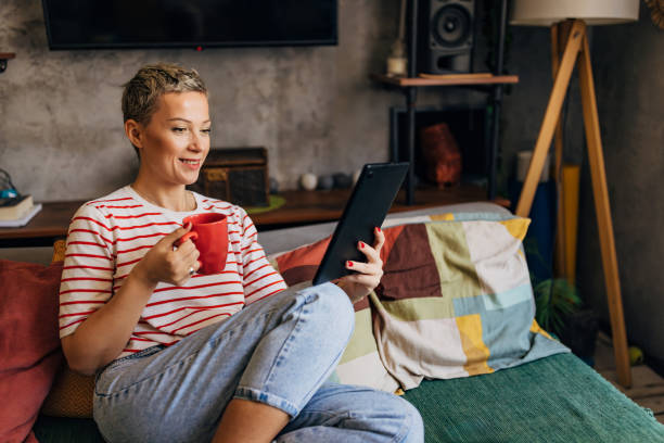 istock Smiling woman is sitting on a comfortable sofa in the living room, drinking coffee and using a digital tablet 1781825032