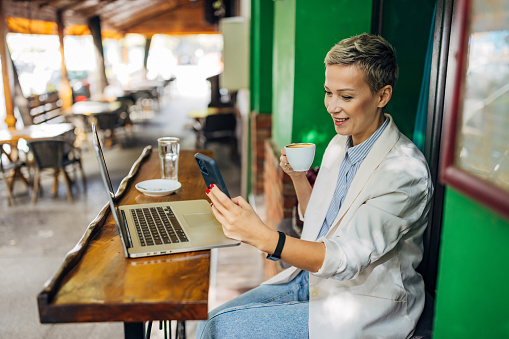 Cheerful businesswoman dressed in casual business attire drinking coffee and using mobile phone and laptop in coffee shop